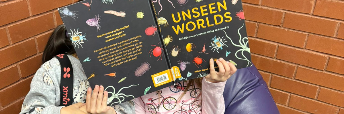Two young readers curled up on a purple beanbag in front of a brick wall, sharing a colourful book titled ‘Unseen Worlds