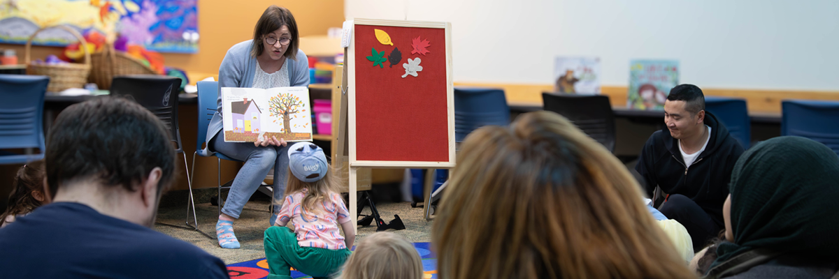 Young child sits on floor in front of Agata, a librarian, who is sitting on chair reading a picture book during storytime. 